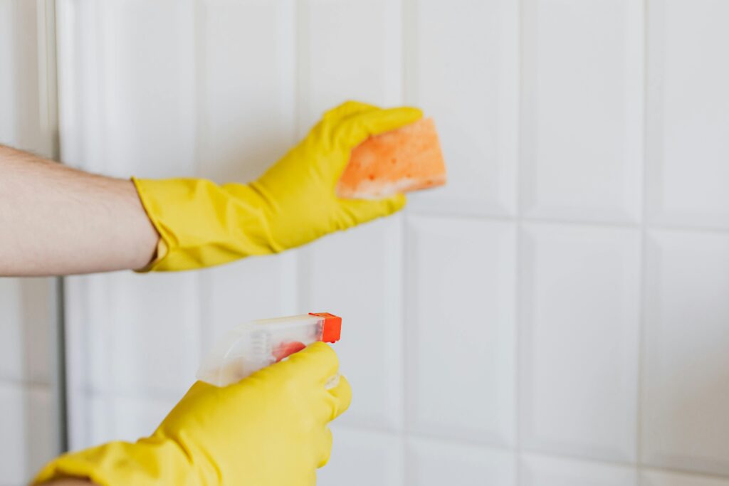 A woman wearing yellow gloves, holding cleaning spray bottles, scrubbing the bathroom wall.