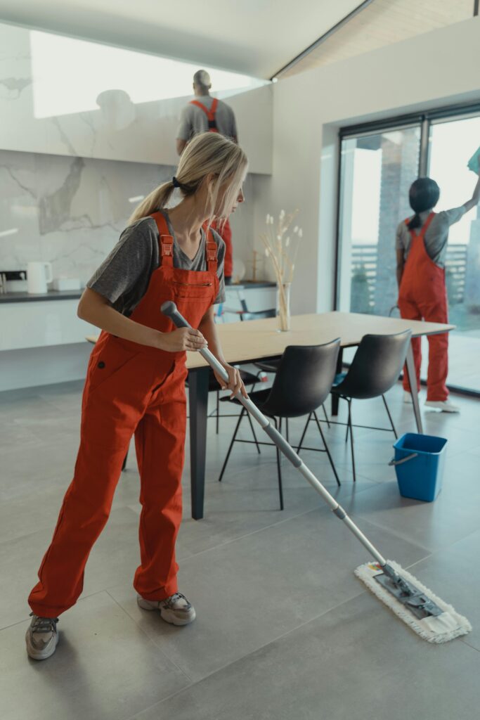 Three people cleaning a spacious living area, mopping the floor and wiping the windows and walls.