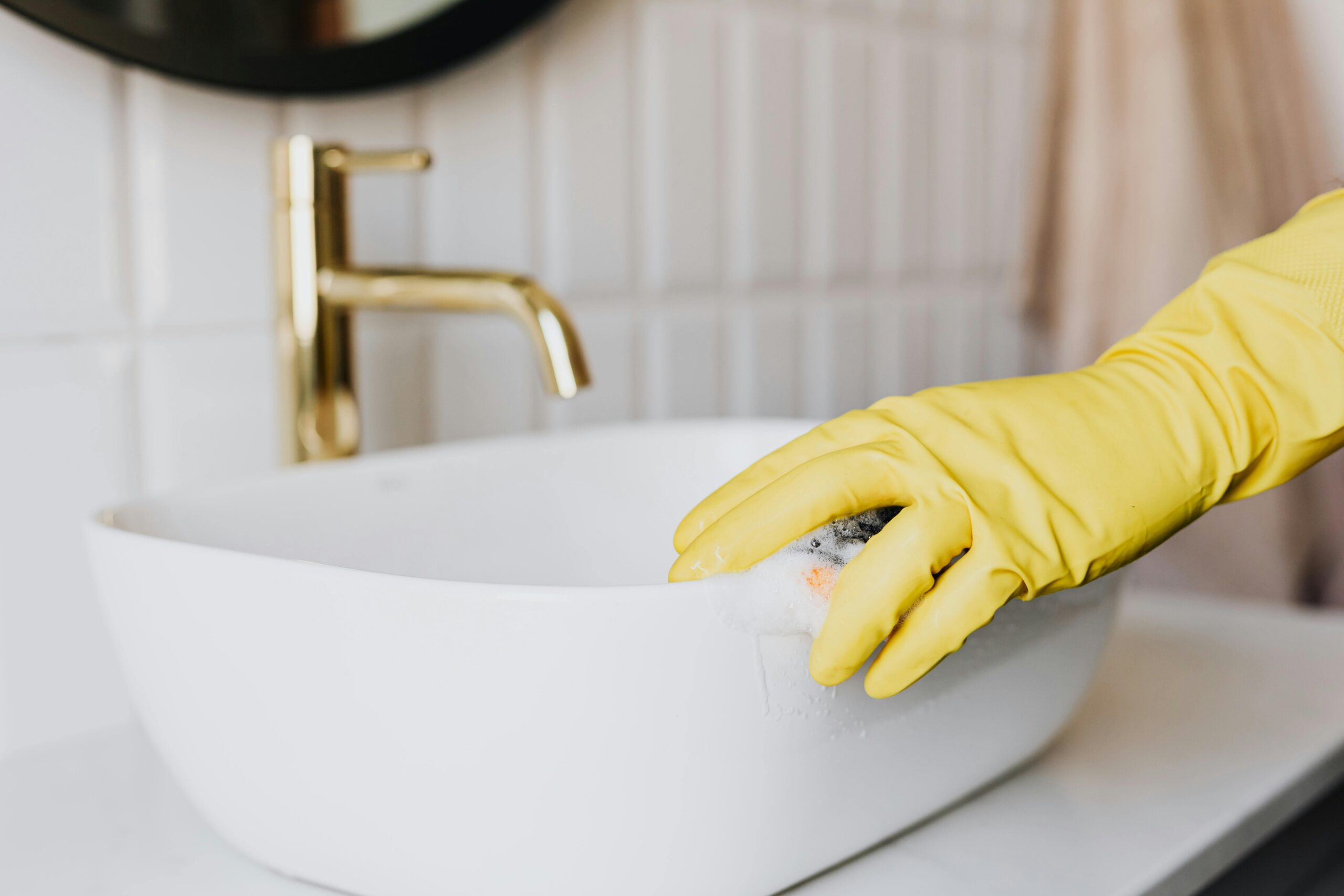 A woman wearing yellow gloves cleaning a white bathroom sink with a gold faucet.